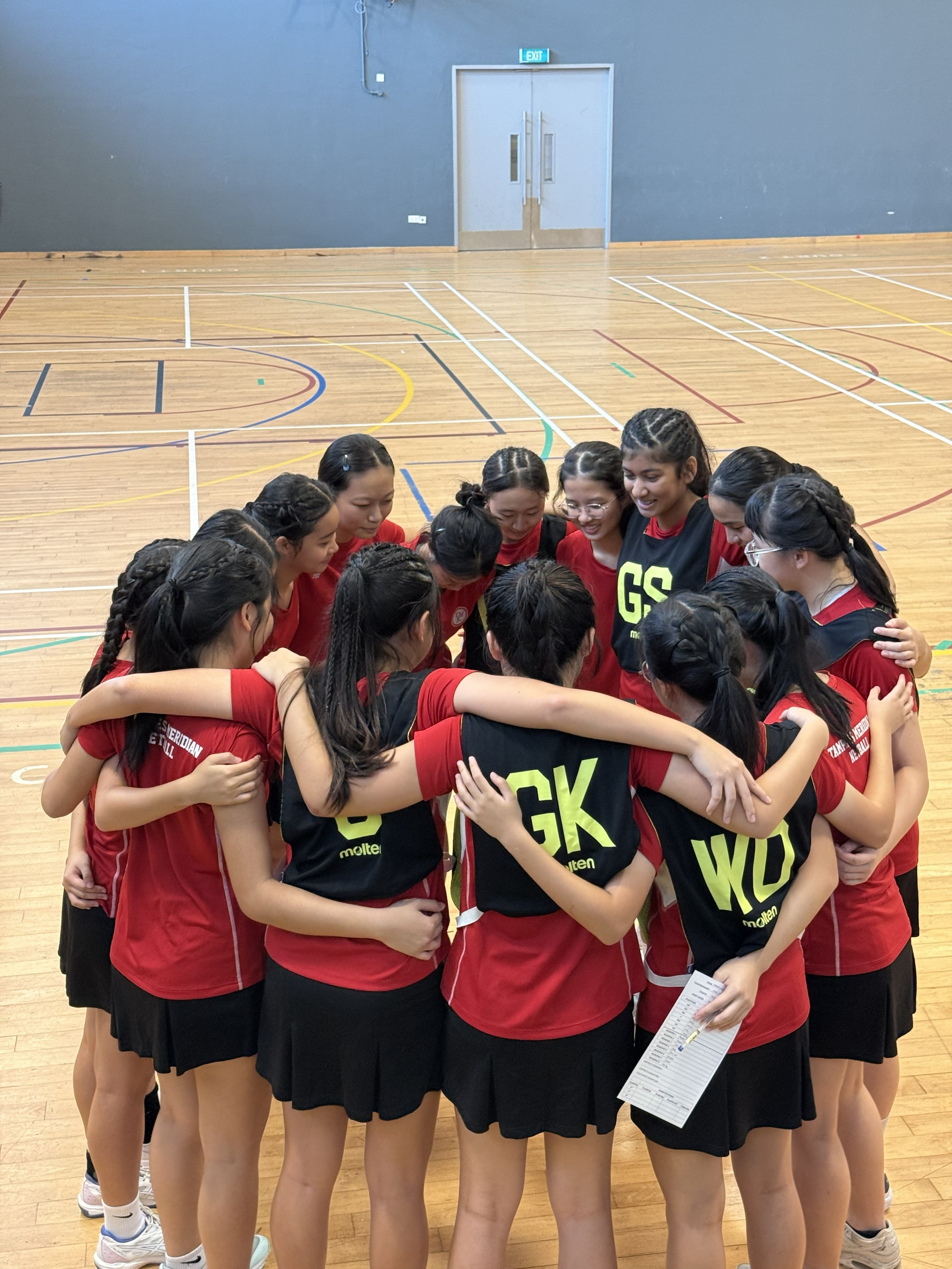 The TMJC netball team huddles together in a circle on an indoor court, arms around each other's shoulders, sharing a pre-match team talk. Players wear red jerseys and black position bibs labelled GK, GS, and WD.